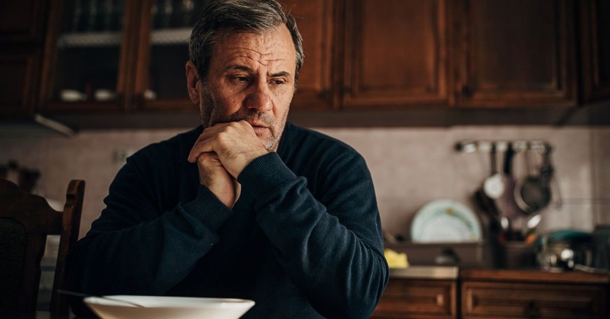 Man sitting at kitchen table with hands folded under his chin and a pensive expression.